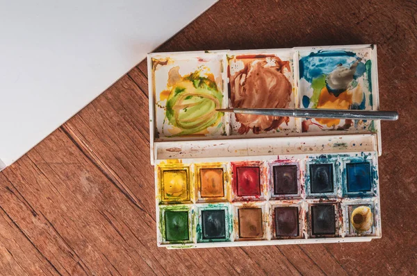 messy box of watercolour paints and brush on wooden table with white paper.