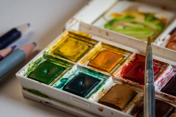 messy box of watercolour paints and brush , close up.with pencils.