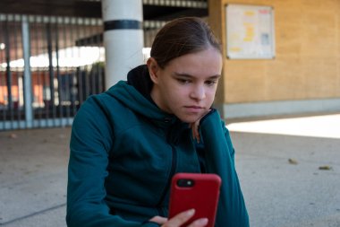 Genç bir kadın dışarıda oturmuş, akıllı telefon videolarına bakıyor. Sıkılmış görünüyor, spor kıyafetleri giyiyor. .