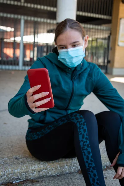 年轻女子戴着面具坐在外面 看智能手机或自拍 穿着运动服 — 图库照片