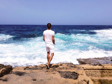 yakışıklı genç adam taşla kenarına beyaz ayakta deniz dalgaları, mavi gökyüzü arıyorsunuz geri, açık seksi manken, sakin, turistik seyahat umuduyla, inanıyorum, çekinmeyin