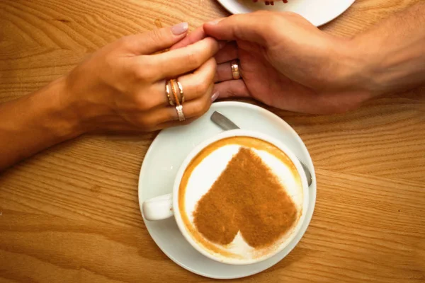 lovely young couple in love sit in a cafe having breakfast,closeup hands holding a cup of coffee with art design heart on it  on a wooden table background