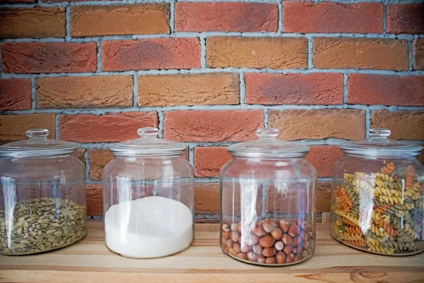 Nueces, pasta, granos y sal en frascos de vidrio en la moderna cocina de diseño loft frente a la pared de ladrillo, concepto de cocina — Foto de Stock