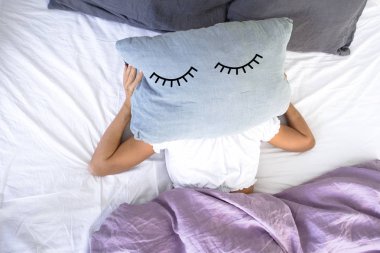 a young girl sleeping in white bed does not want to get up early in the morning,covering her face with pillow with closed eyes on it. clipart