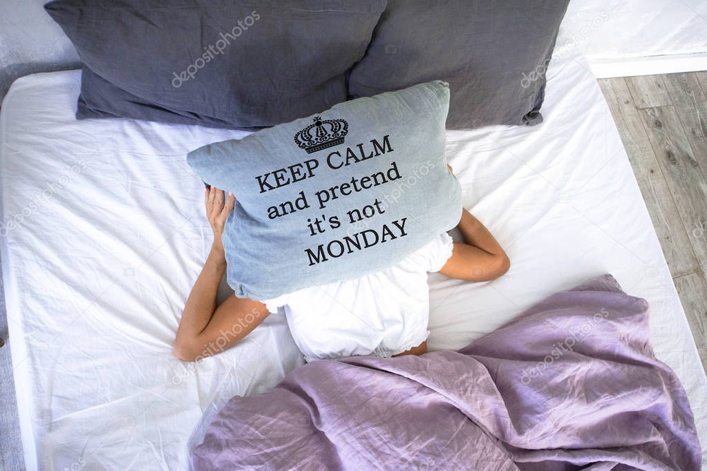 a young girl sleeping in white bed doesn't want to get up early in the morning,covering her face with pillow with words keep calm and pretend it is not Monday.