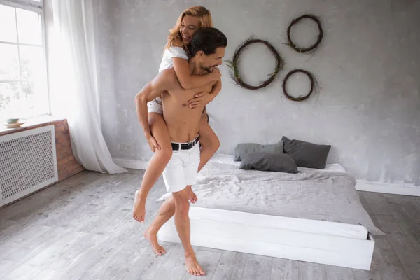 若く美しく新しいモダンなベッドルームで一緒に楽しんで幸せなカップル男の愛の物語、彼の背中に彼のガール フレンドを運ぶ — ストック写真