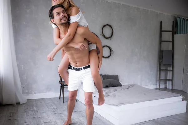 Una giovane bella coppia felice divertirsi insieme in una nuova camera da letto moderna, uomo portando la sua ragazza sulla schiena, storia d'amore — Foto Stock