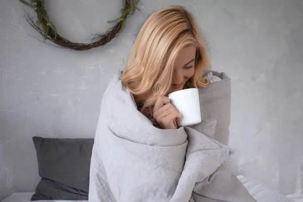 Seorang gadis cantik duduk di tempat tidur di hari yang dingin menutupi dengan penutup dan minum teh, mencari dan tersenyum — Stok Foto