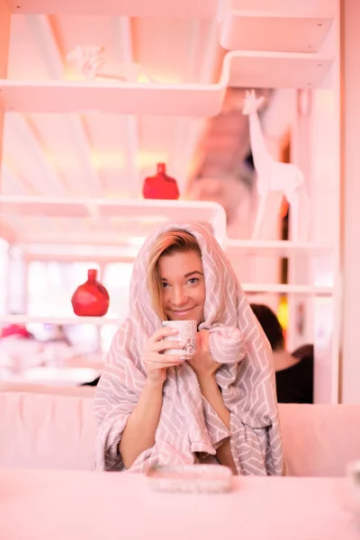 Μια όμορφη νεαρή κοπέλα που κάθεται σε ένα καφέ με ένα κάλυμμα στο κεφάλι της, όπως αυτή είναι κρύο χαμογελώντας και πίνοντας ζεστό τσάι που προσπαθεί να πάρει ζεστό — Φωτογραφία Αρχείου