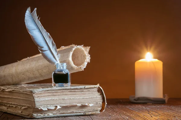 Quill pen, inkwell and a rolled papyrus sheet on an old book by candle light