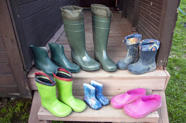 Old dirty rubber boots of a big family stand on the wooden floor of a country house porch lit by summer sun with green grass. Short rubber country goloshes.