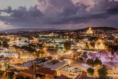 Gürcistan 'ın başkenti Tiflis' in güzel panoramik manzarası. Şehir rengarenk ışıkları, nehir, katedral, arka plandaki güzel günbatımı dağlarındaki eski binalar. Akşam manzarası.