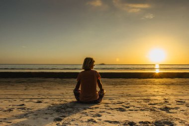 Gün batımında sahilde oturan genç bir kızın silueti. Bulutlar, deniz ve kumlu sahil. Deniz kenarında romantik akşam meditasyonu. Yaz tatili. Renkli su yansıması. Tropikal cennette rahatla.