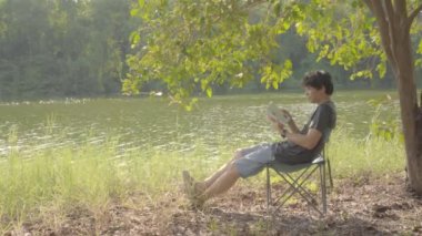 4K vdo adamı yazın gölün kenarında mutlu bir şekilde kitap okurken doğada dinleniyor.