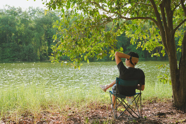 relax and lifestyle activity from asian man 30s to 40s during sit and put arm on head at side of river and under tree on spring season with happiness feeling with sunset background