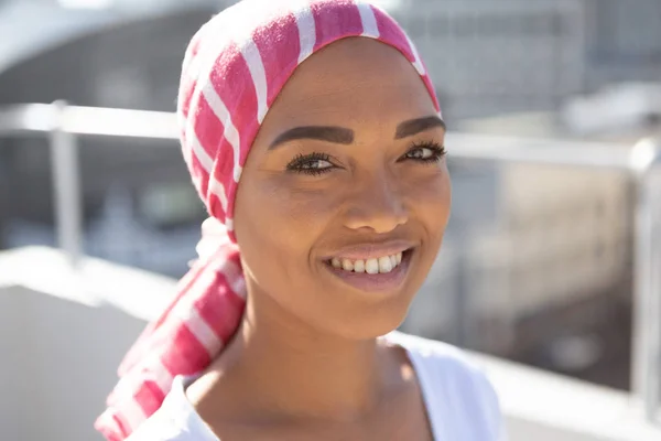 Portrait of smiling woman wearing scarf with breast cancer awareness against city background