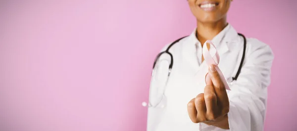 ピンクの背景の乳がん啓発ピンクリボンを保持している笑顔の看護師 — ストック写真
