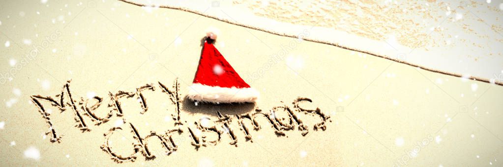 Snow falling against merry christmas written on sand with santa hat