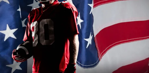 アメリカの国旗のクローズ アップに対してラグビー ボールを保持しているヘルメットのアメリカン フットボール プレーヤー — ストック写真
