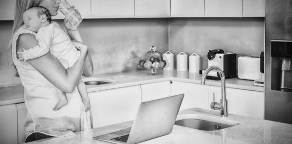 Stock image Young woman drinking water while carrying baby in kitchen at home