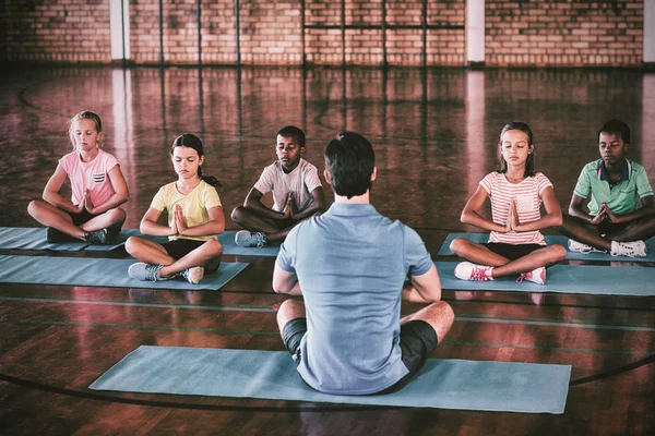 School kids and teacher meditating during yoga class in basketball court at school gym