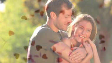 Yakışıklı beyaz erkek kucaklayan güzel beyaz kadın arkadan küçük kalpler ön planda park devam ederken dijital bileşik. Çift gülerek ve gülümseyerek