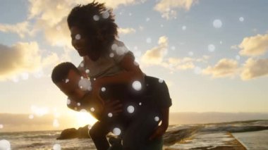 Close of a couple where the Caucasian man carries the African-american woman on his back while walking on the shore at sunset
