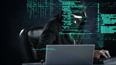 Siyah gözlük takan sinsi bir hacker ve bilgisayarda yazan bir kapüşonlunun yan görünümü. Arka plan hareketli arabirim kodları ile doldurulur