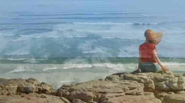 Sahilde kayaların üzerinde oturan kızıl saçlı kadının arka görünümü