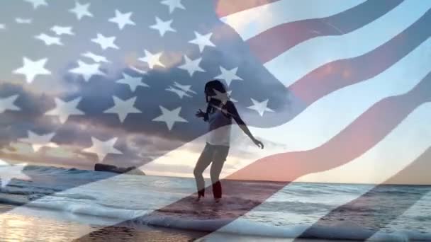 前景で手を振るアメリカの旗を持つビーチで波で遊ぶ女性のデジタルコンポジット — ストック動画