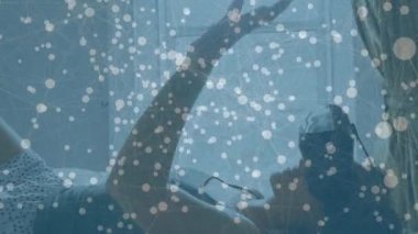 Ön planda bağlı çizgiler ve noktalar ile bir sanal gerçeklik kulaklık takarken yatakta yatan bir kadının dijital kompozit