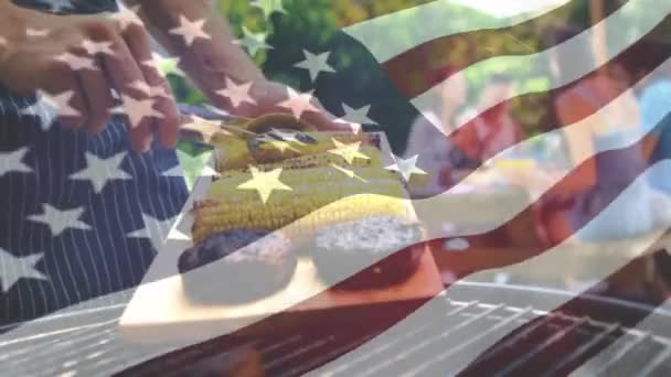 一个人烤玉米和肉 一面美国国旗飘扬在前景的数字合成 他身后是他的朋友们 他们坐在一张野餐桌旁 — 图库视频影像