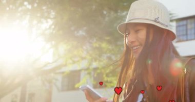 Kova şapka ve deri ceket giyen Asyalı bir kadının güneş parlarken cep telefonuna bakarken gülerek gülmesinin dijital bileşimi güneş parlarken ve dijital kalpler yukarı doğru 4k hareket ederken