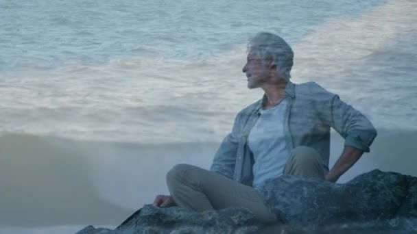 岩の上に座って 日没時に海の背景に集中する古い白人男性のデジタルコンポジット — ストック動画