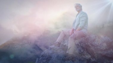 Bulutlu bir arka plan ve güneş ışınları ile bir kaya üzerinde otururken sakin görünümlü eski bir Kafkas adam dijital kompozit