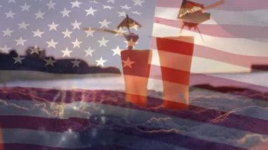 Gün batımında plajda kuma yerleştirilen gözlüklerdeki iki kokteylin dijital bileşimi, bokeh ışıklı 4 Temmuz metninin arkasında Amerikan bayrağı dalgadalga gelirken.