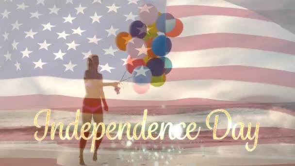 7月4日のボケライトで金の独立記念日のテキストの後ろにアメリカの旗が波立つ間 ビーチで風船を持つ白人女性のデジタルコンポジット — ストック動画