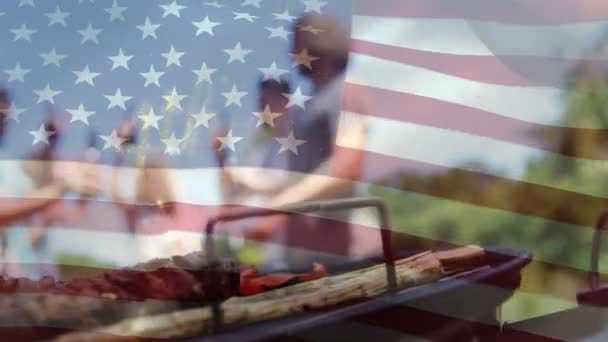 ボケライト付きの7月4日のゴールドテキストのデジタルアニメーションは アメリカの旗がバックグラウンドで波立っている間に表示されます 様々な友人のグループは 飲み物やバーベキューを通して屋外で祝います — ストック動画