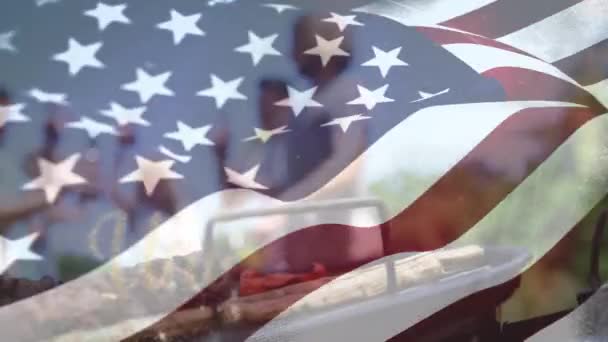 背景に手を振っているアメリカの旗の前に現れる筆記体の金の米国のテキストのデジタルアニメーションは 7月4日のために飲み物やバーベキューを祝う多様な人々のグループを示しています — ストック動画