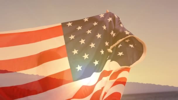 在海滩边举着美国国旗的人的数字合成 而背景显示日落 — 图库视频影像