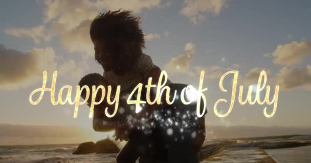 日没時にビーチでアフリカ系アメリカ人のカップルのデジタルコンポジットが 白い光沢のあるライトで7月4日の金ハッピー4日の挨拶が前景に表示されます — ストック動画