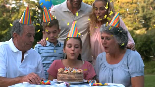 屋外で若い女の子の誕生日を祝う3世代の白人家族のアニメーション 誕生日ケーキにろうそくを吹き出し パーティーの帽子を身に着けています コンフェッティは前景に落ちている間 — ストック動画