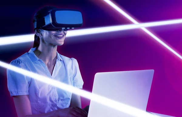 女性はターコイズと紫の背景に対してコンピュータ上でバーチャルリアリティヘッドセットを使用します — ストック写真