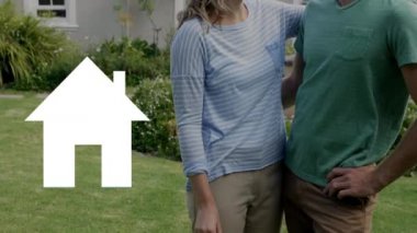 Evlerinin bahçesinde dikilen mutlu beyaz çiftin eğik çekimleri. Evin bir simgesi mavi ile doluyken elinde anahtarlar olan kadın.