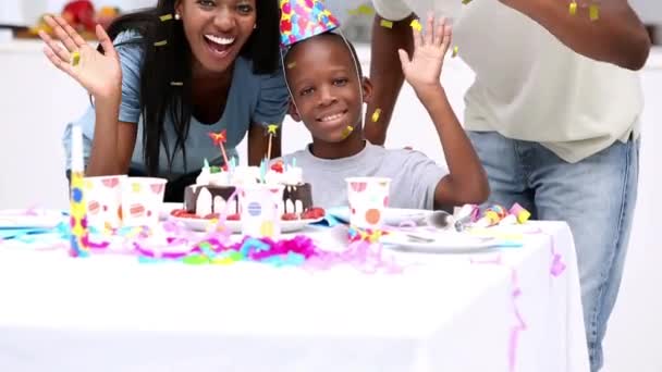 誕生日パーティーで息子と一緒に若いアフリカ系アメリカ人のカップルのアニメーションは カメラに手を振って笑顔とパーティーの帽子を身に着けていますが 黄金のコンフェッティは前景に落ちています — ストック動画