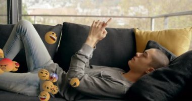 Arkadan sağa sola uçan emoji simgelerinin animasyonu, arka planda yatan ve akıllı telefon kullanan genç bir beyaz adamın yan görüntüsü.