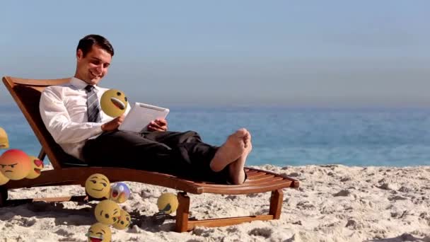 左から右へ飛ぶ絵文字のアイコンのアニメーション 背景にあるビーチでタブレットを使用している若い白人男性 — ストック動画