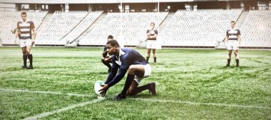 Afrika kökenli Amerikalı erkek rugby oyuncusu stadyumdaki bir standa rugby topu yerleştiriyor. Arka planda oyuncular.