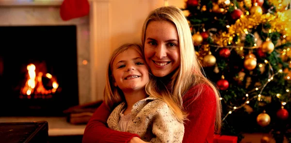 若い白人女性が 飾られたクリスマスツリーのそばのクリスマスの時に居間で幼い娘を膝に抱きしめ カメラに微笑む正面図 — ストック写真