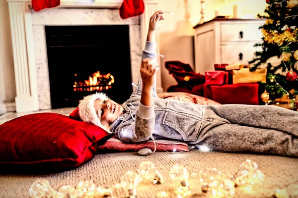 圣诞节时 一个快乐的白人小男孩躺在地板上 戴着圣诞老人的帽子 在客厅里拿着仙女灯 — 图库照片
